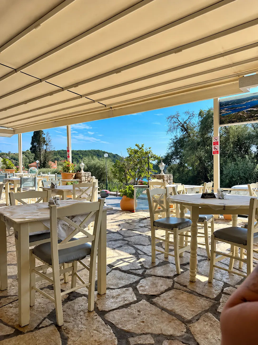 Outdoor seating area of O Meraklis taverna with tables under a canopy overlooking the hills of Paleokastritsa
